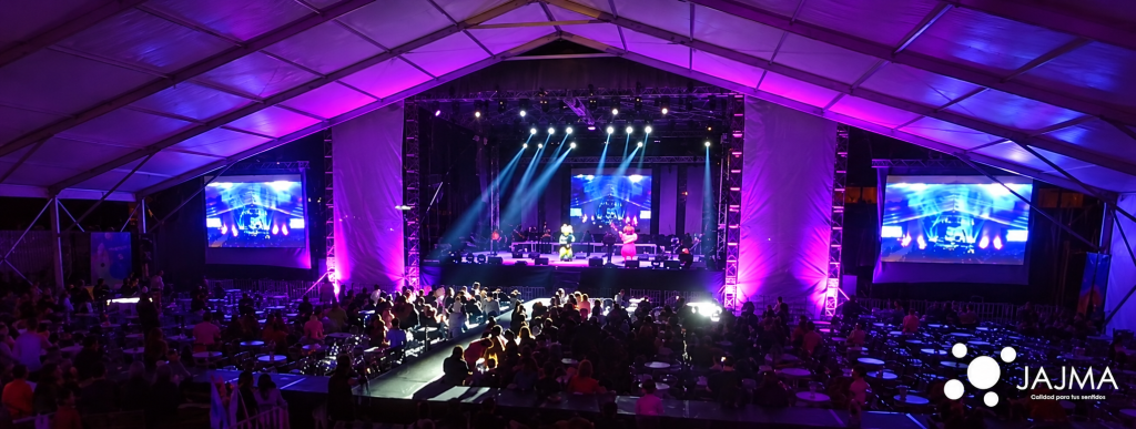 Vista panorámica del evento bajo carpa con pantallas e iluminación morada.